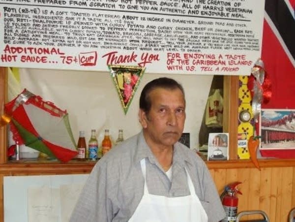 A man in a white apron poses for a photo under a restaurant menu.