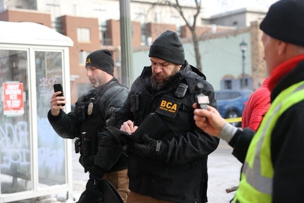 Investigators in black coats take notes and photos on a phone.