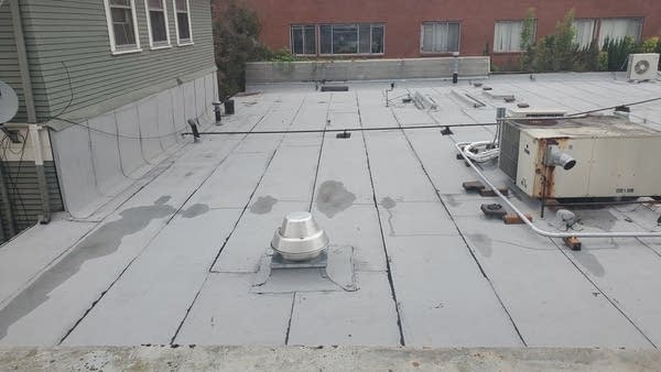 Top of a flat, gray roof w/ air conditioning unit and puddles of water