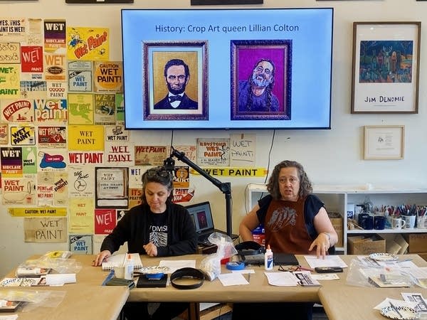 Two women sit at a table. The wall behind them is covered in crop art pieces, and the screen above them shows more crop art.
