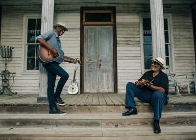 Two men play guitars on the front porch of a house