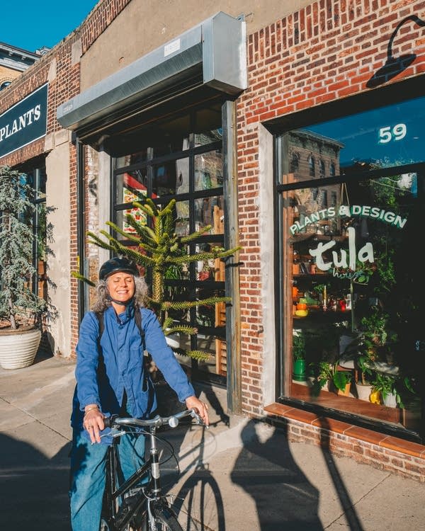 A woman with gray hair on a bicycle smiles in the sun outside of a Brooklyn plant shop.