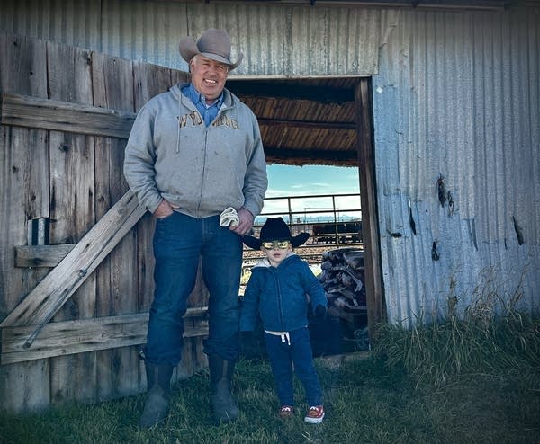 A man in a tan cowboy hat and toddler in a black cowboy hat. They are standing in front of a wooden barn door.