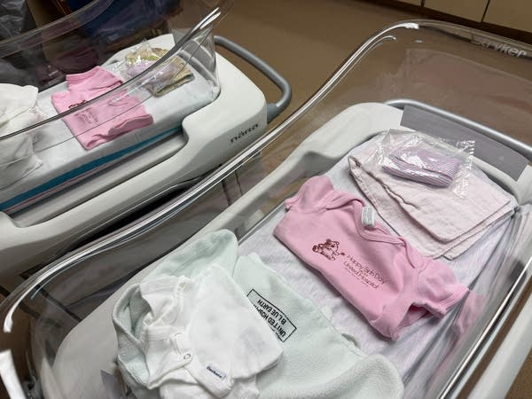 Two hospital cribs have baby clothes inside them.