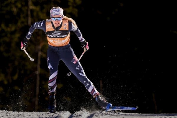 A woman in an orange and black suit skis down a hill.