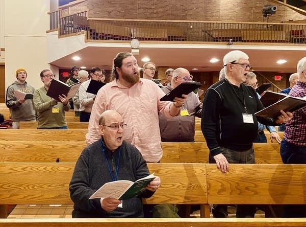 A man sits in a pew, others behind him stand as they all sing.
