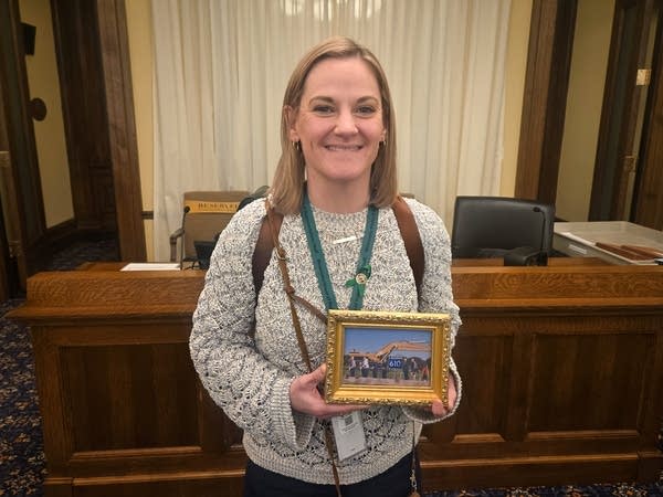 A woman in an office holds a golden framed photo of people breaking ground on a highway.