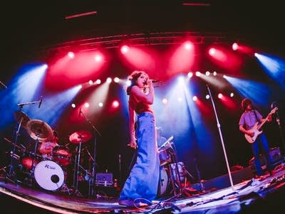 A woman sings into a microphone on a lighted stage with a band behind her.