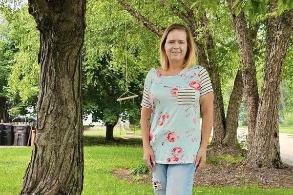 Elizabeth Amirault wears a light blue, green and pink floral shirt and blue jeans. She is standing outside, near trees.