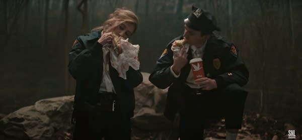 TV Image of two cops eating hoagies at crime scene in the woods