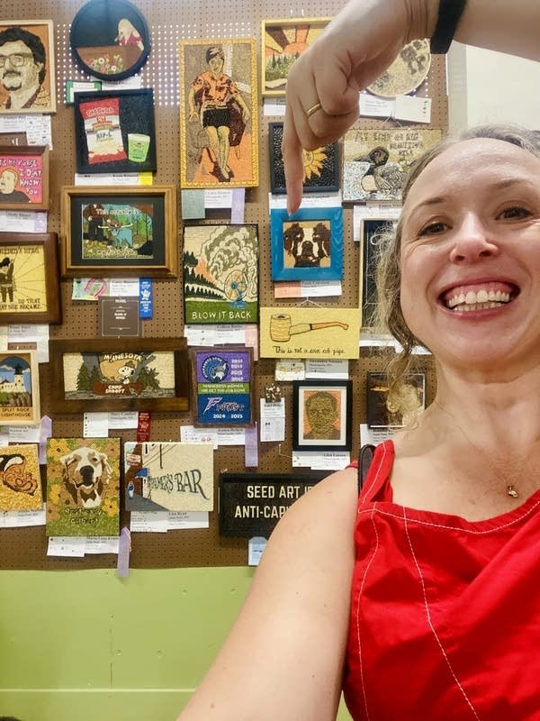 A woman in a red shirt grins in a selfie while pointing to her crop art in the background.