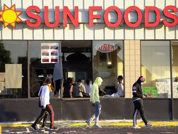 People walk by a storefront with broken glass, with more people inside.