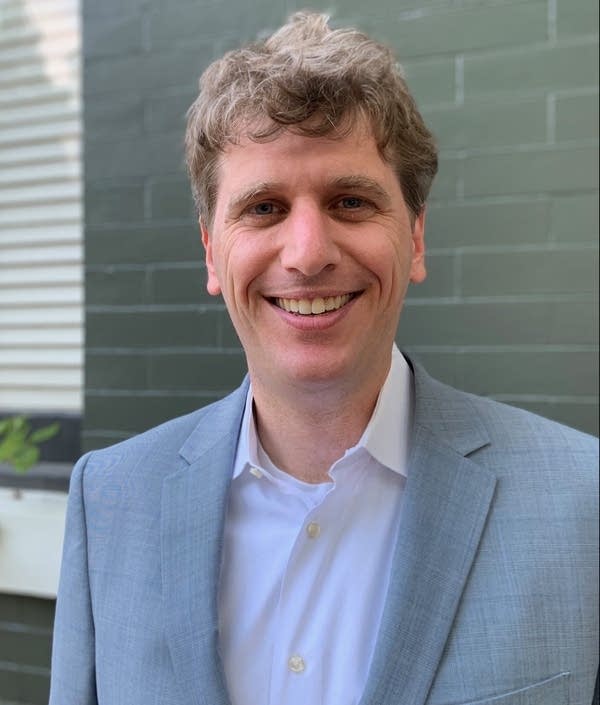 Matt Stoller smiles for his headshot as he stands in front of a grey brick background.