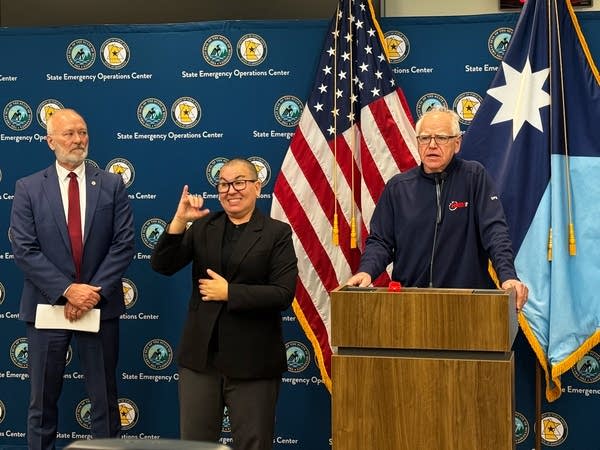 A man in a zip-up jacket speaks at a podium while an ASL interpreter signs next to him.