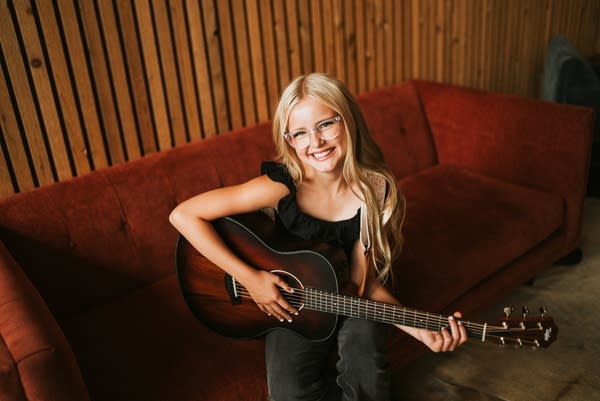 Brooklyn Marie poses at a red couch with her guitar.