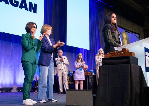 Two women clap while another woman speaks at a podium on stage.