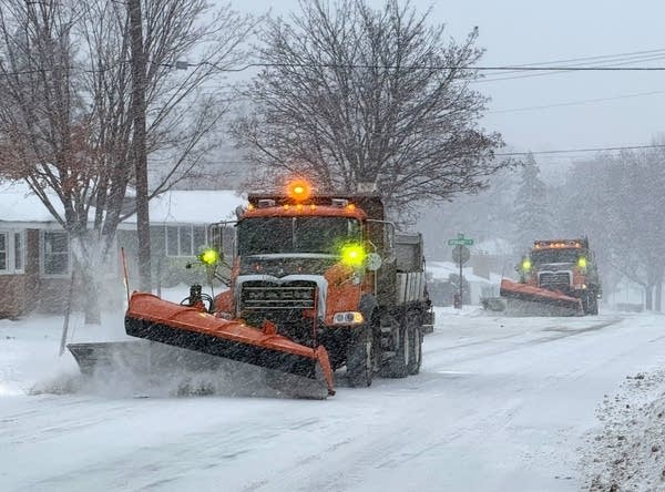 Two snow plows drive down a residential street during a snow storm.
