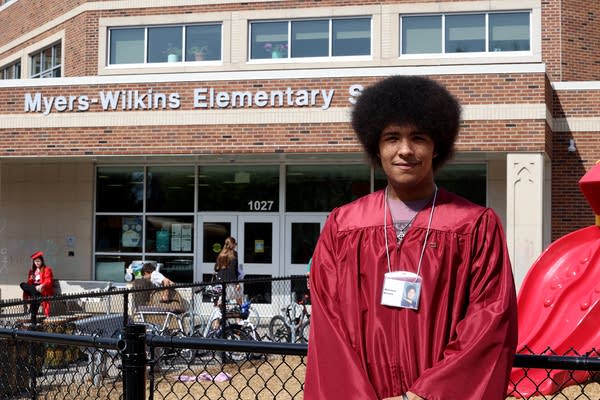 A student stands in a red graduation robe outside of a school.