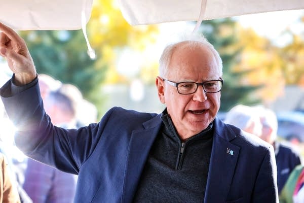 A man in a suit and glasses raises his hand while speaking.