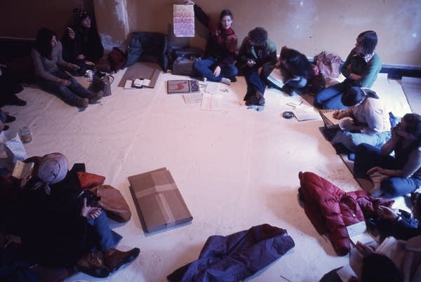 A large group gathers in a room, sitting on the floor.