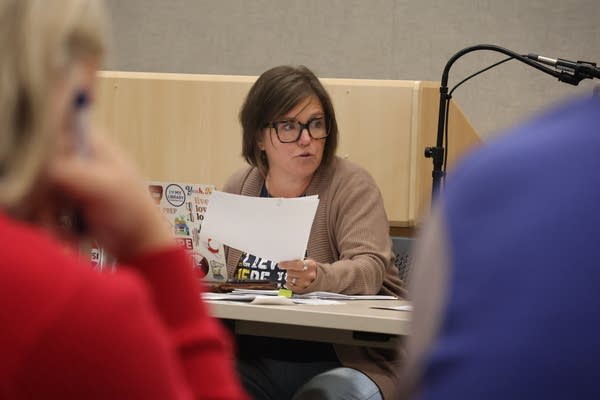 A woman sitting at a table talks to someone just offscreen.