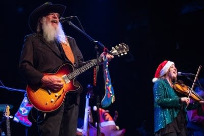 A bearded man sings and plays guitar onstage; a fiddle player accompanies