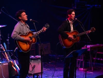 Two men sing into microphones and play acoustic guitars.