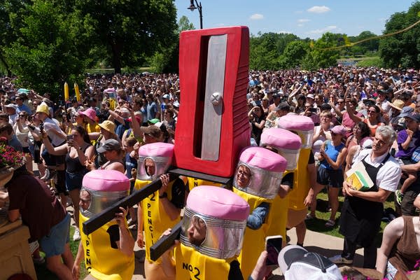 Six people in pencil costumes carry a giant red pencil sharpener like pall bearers.