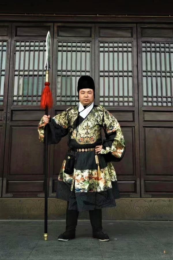 Cheng Shi is dressed up in traditional Chinese costume. It is multicolored and he is holding a large staff.