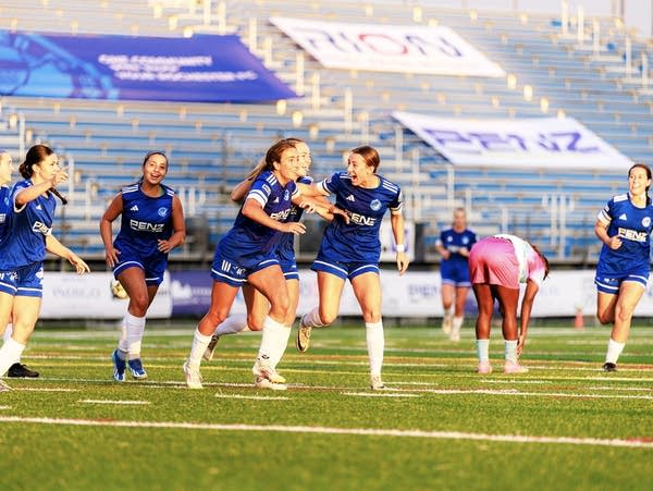A group of soccer players wearing blue uniforms run smiling on their home field.