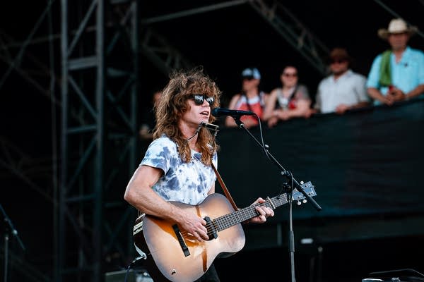 A musician sings and plays guitar on an outdoor stage