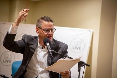 A man holds one arm up while speaking into a microphone and looking at papers in his other hand.