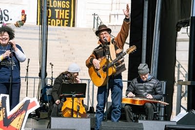 A musician sings and plays guitar on an outdoor stage at a large rally