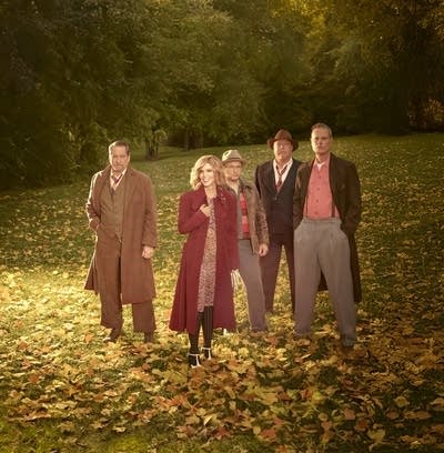 Five people in long coats standing in a field of autumn leaves
