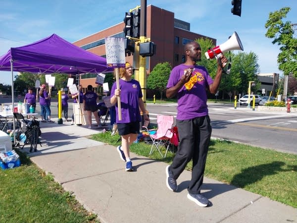 Protesters march on a picket line in purple, one holds a megaphone, another holds a sign.