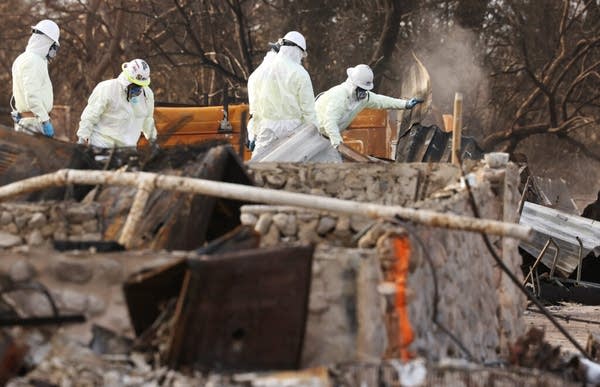 People in white hazmat suits pick through the debris of a charred home.