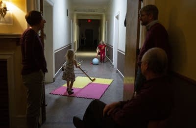 Two children play with a ball and hockey sticks in a hallway.