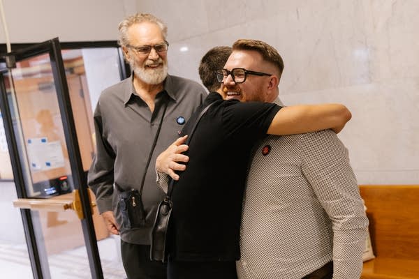 Two people hugging while another person smiles in background