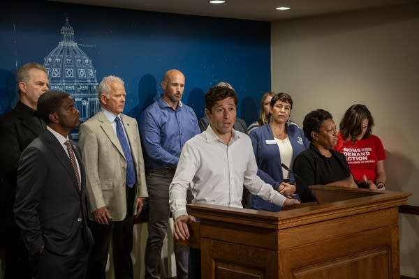 A man in a white shirt speaks at a podiium surrounded by people in a small conference room.