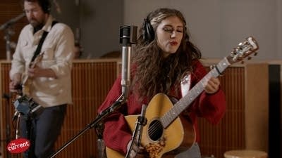 A woman sings and plays guitar and a man plays guitar in a recording studio