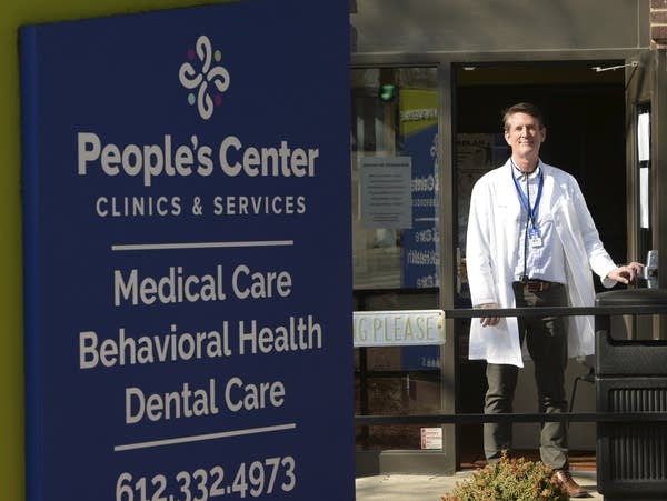 A man in a white coat stands outside the door of a clinic.