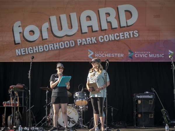 An average-height white woman holds a microphone and a clipboard on stage