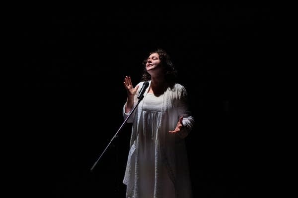 A woman stands on a dark stage with a mic in front of her.