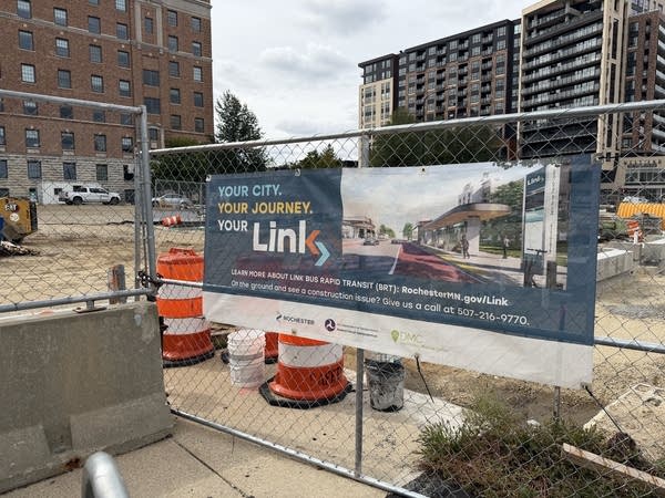 Signs and construction cones sit near a road construction site, fencing surrounds the site.