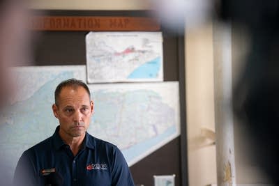 A man in a dark blue polo shirt stands in front of a wall of maps.