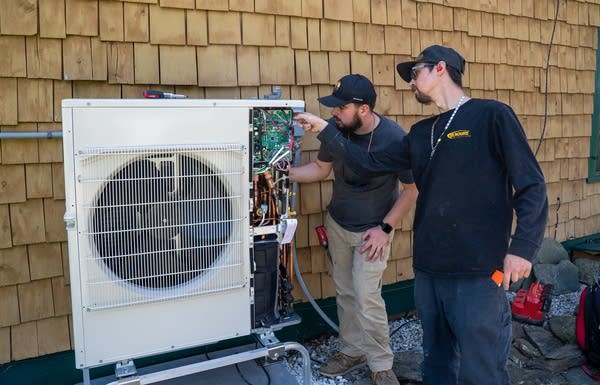 Two men install an HVAC unit for a heat pump on the side of a house.