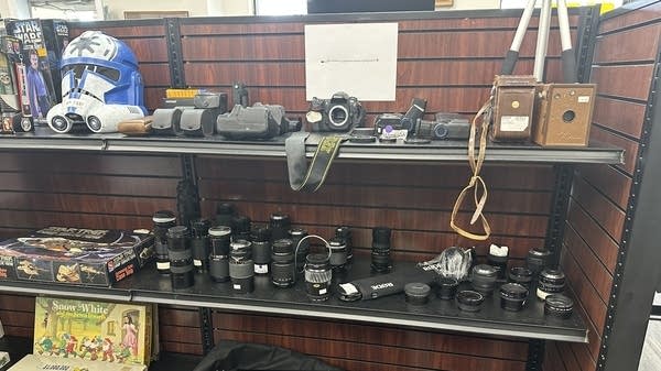Three shelves hold a variety of items: a Stormtrooper helmet, some camera bodies, a whole row of camera lenses of different sizes, a tripod, and some board games.