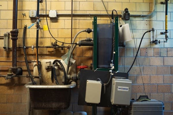 A sink, pipes and a large tank hang on a brick wall.