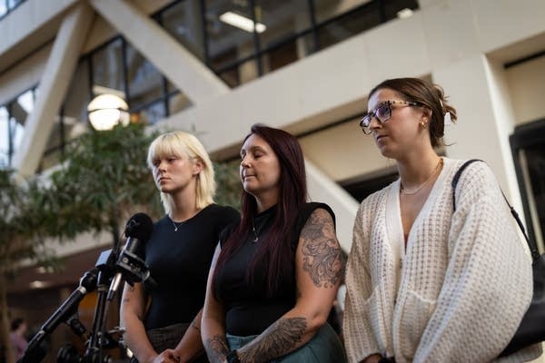 Three women stand, one speaks to the press