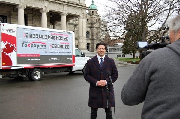 Franco Terrazzano, a man in a suit and tie with a dark overcoat and black hair, poses in front of a camera man with a TV camera wearing a gray sweatshirt. He is standing on pavement in front of a large truck sponsored by the Canadian Taxpayer Federation that lists the Canadian Federal Debt.
